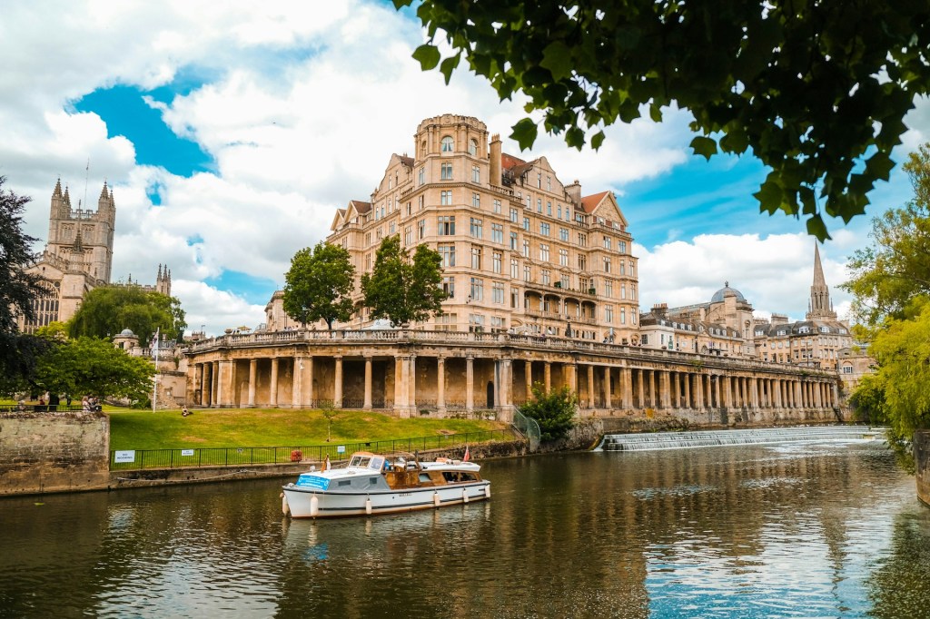 A boat motors along a river with an ornate building behind in Bath.