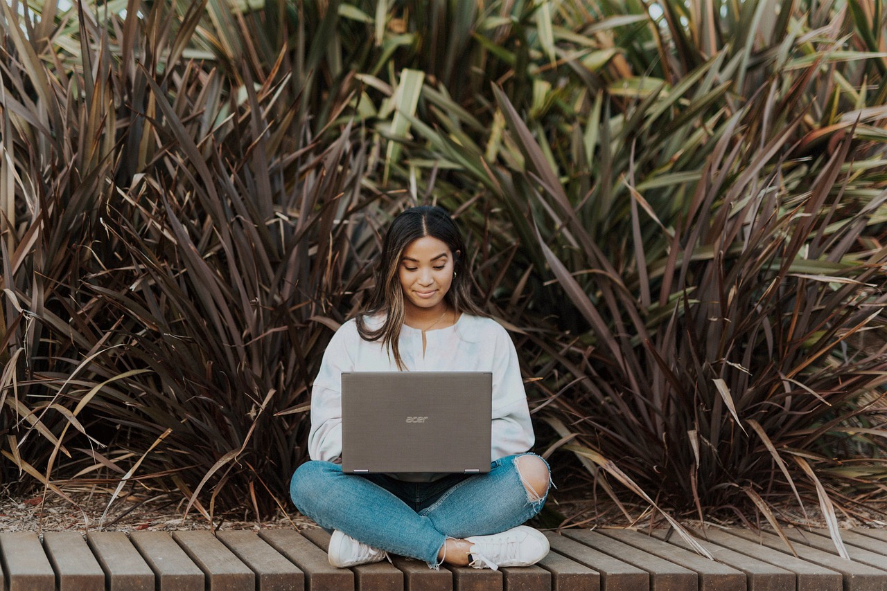 A woman working on a laptop sitting on a boardwalk outdoors.