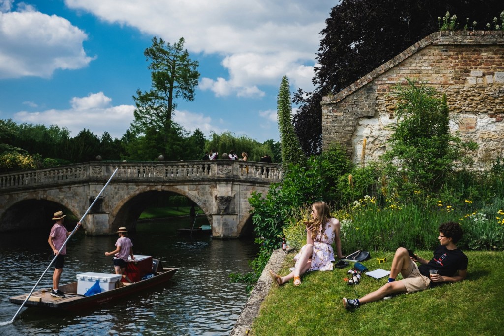 People sitting next to the river in Cambridge as a punt goes by.