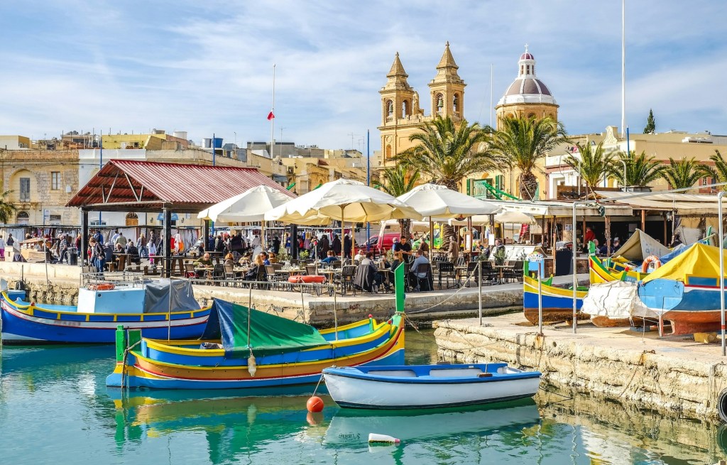 A busy scene on the harbour side in Malta with people sitting at tables under canvas covers.