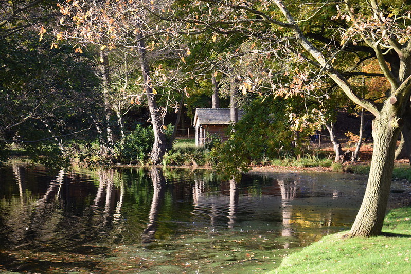 A view across a lake to a small stone building surrounded by trees.