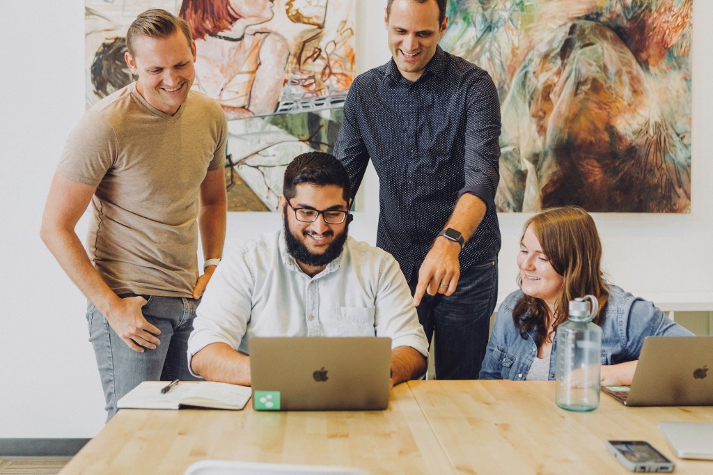 A group of co-workers gathered around a laptop laughing and talking.