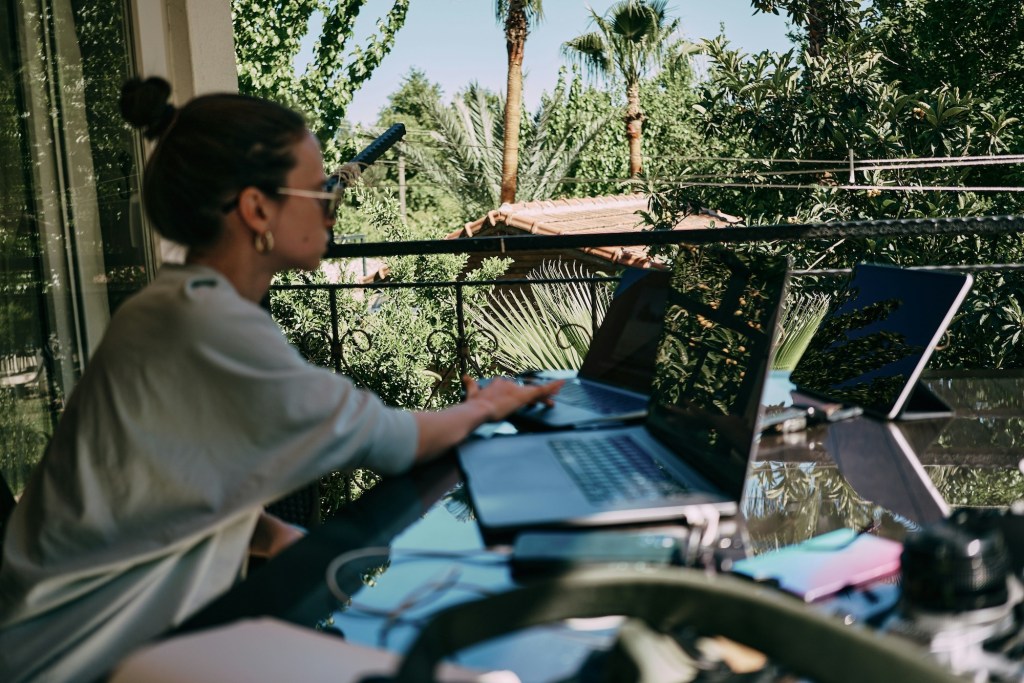 A young woman works at a laptop on a balcony in an exotic location.